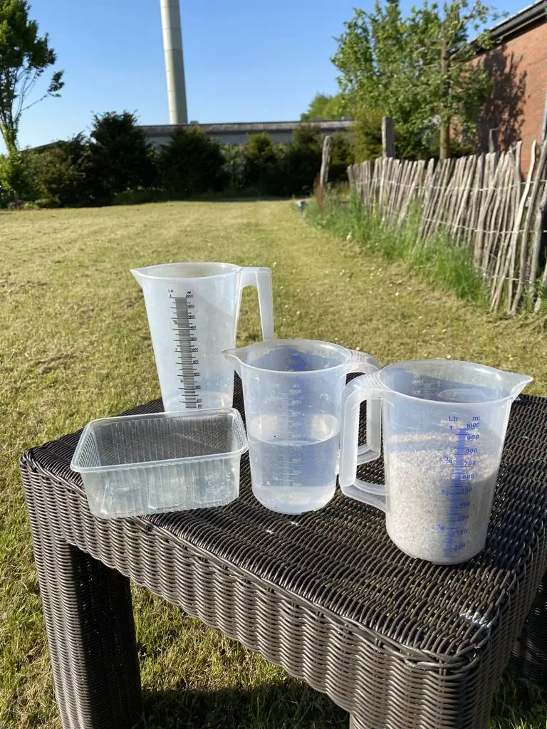 Drei Messbecher und eine Schale auf einem Gartentisch, einer mit Wasser, einer mit hellem Granulat